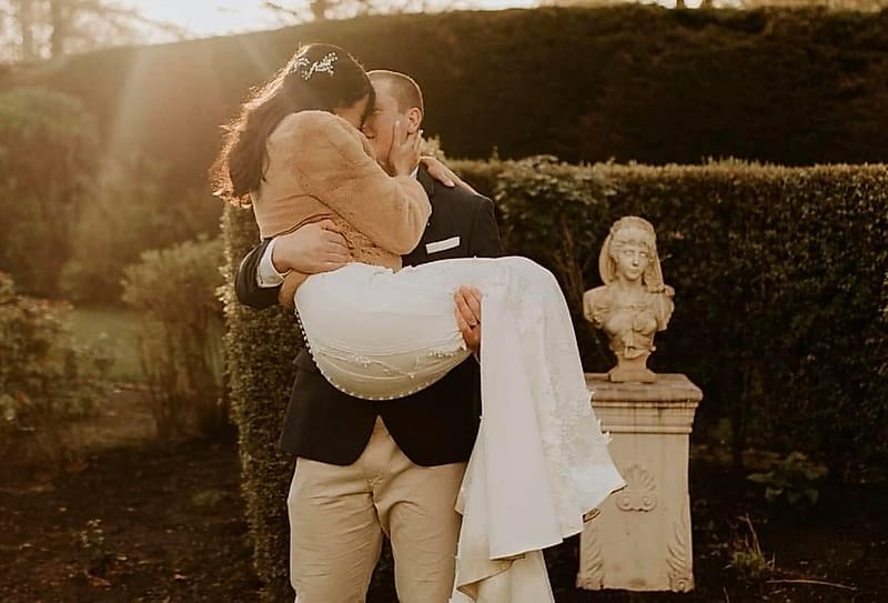 Groom carries the bride in golden evening light; her gown has covered buttons down the back and beaded lace on the skirt