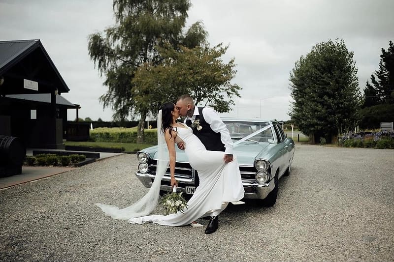 Groom dips the bride beside a vintage car; she wears a fitted crepe gown and cathedral veil