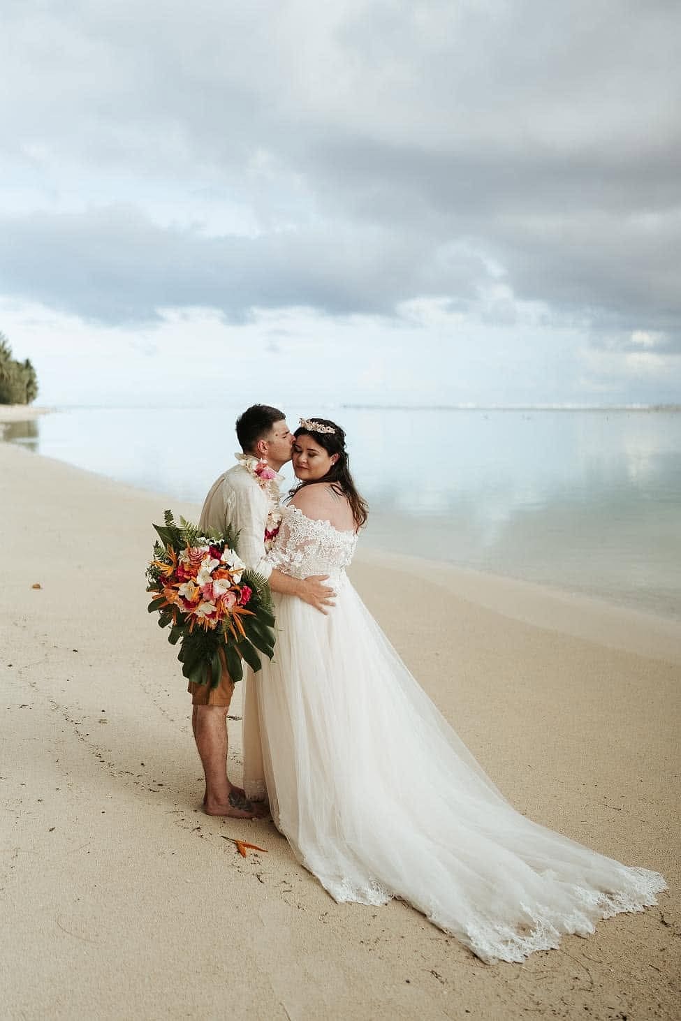 Bride and groom on a beach; her gown has off-the-shoulder lace sleeves and a full tulle skirt