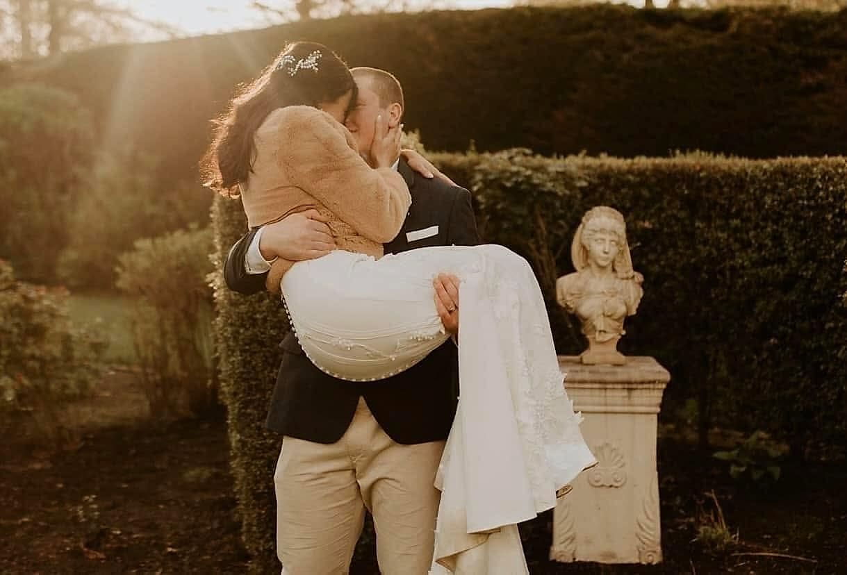 Groom carries the bride in golden evening light; her gown has covered buttons down the back and beaded lace on the skirt
