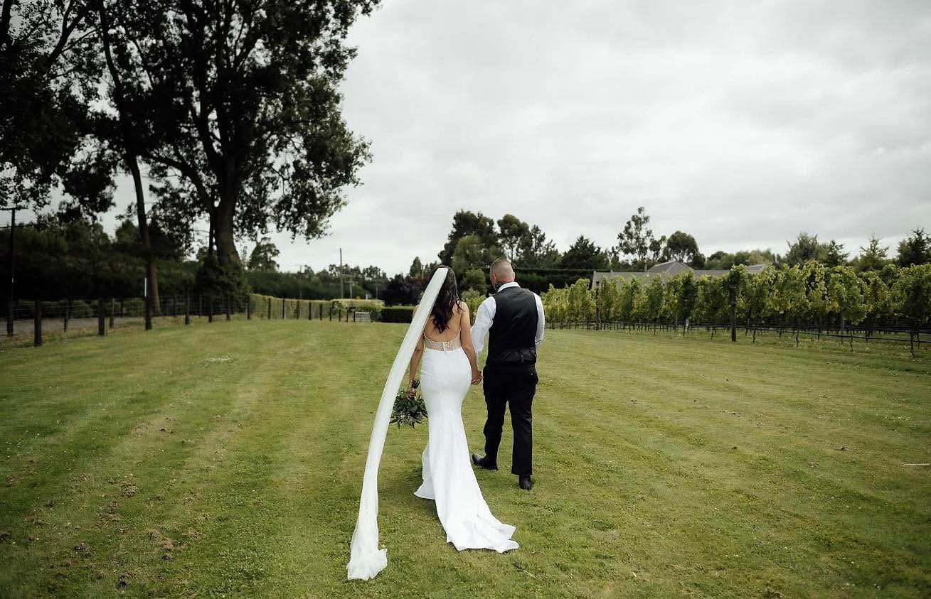 Bride and groom walk through a vineyard as her long veil lifts in the wind