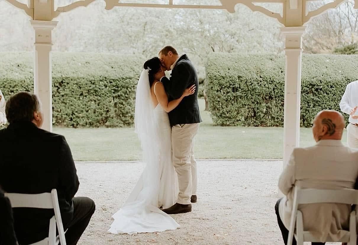 Bride and groom kiss under a white gazebo; she wears a fitted gown and long veil